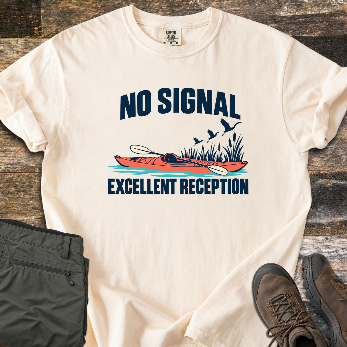 Excellent Reception Comfort Colors T-shirt in Ivory, laid flat. The words No Signal arch over an orange kayak with a paddle among reeds and flying birds, above the words Excellent Reception. Olive pants and brown hiking boots sit on a weathered wood surface.