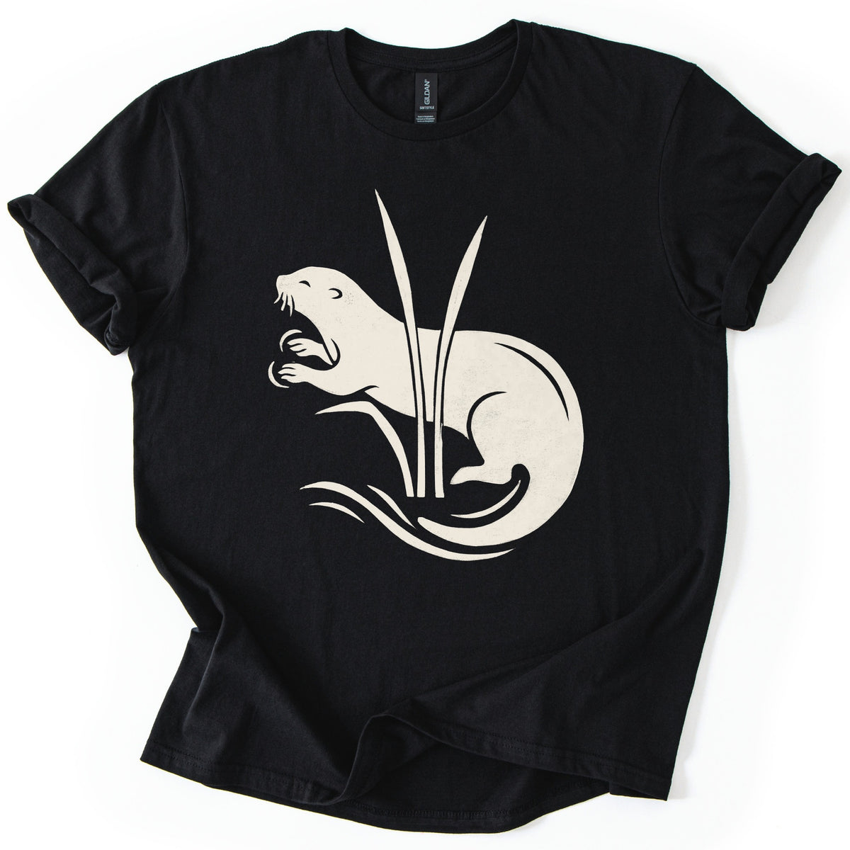 Otter In The Reeds Comfort Colors T-shirt in Black, laid flat. A cream silhouette of an otter with open mouth, whiskers and raised paws curls behind two tall reeds above swirling water lines. No words. The shirt lies on a white surface with sleeves rolled.