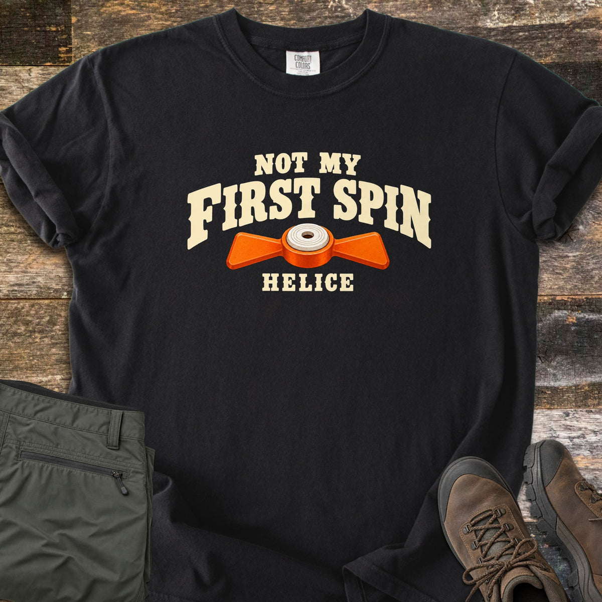 Not My First Spin Comfort Colors T-shirt in Black, laid flat. Cream arched text reads Not My First Spin above an orange two-winged helice target with a white center, with Helice below. Folded olive pants and brown hiking boots sit beside it on weathered wood planks.