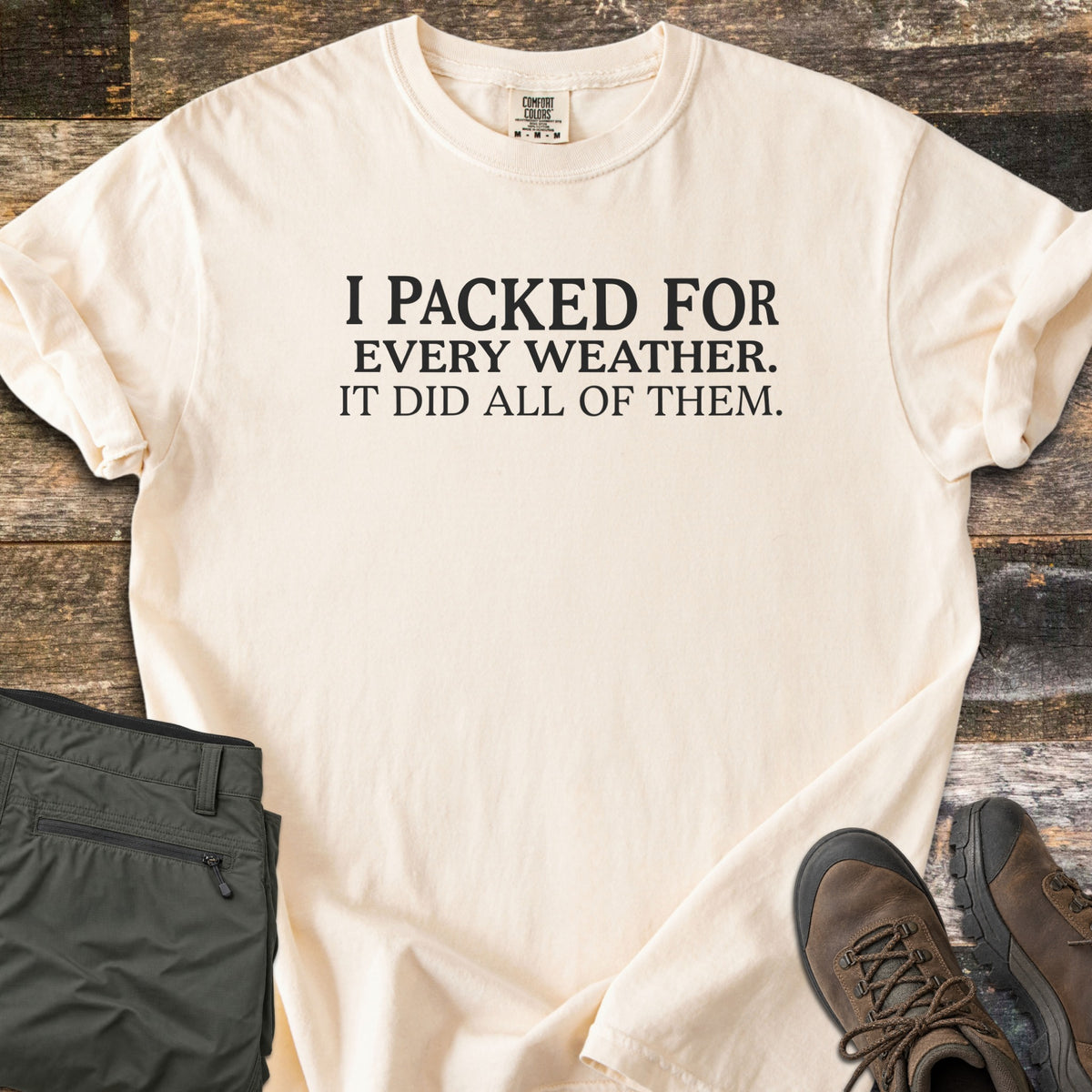 Packed For Every Weather Comfort Colors T-shirt in Ivory, laid flat. Black serif text on the chest reads I PACKED FOR EVERY WEATHER. IT DID ALL OF THEM. The shirt lies on weathered wood planks with folded olive hiking pants and brown hiking boots.