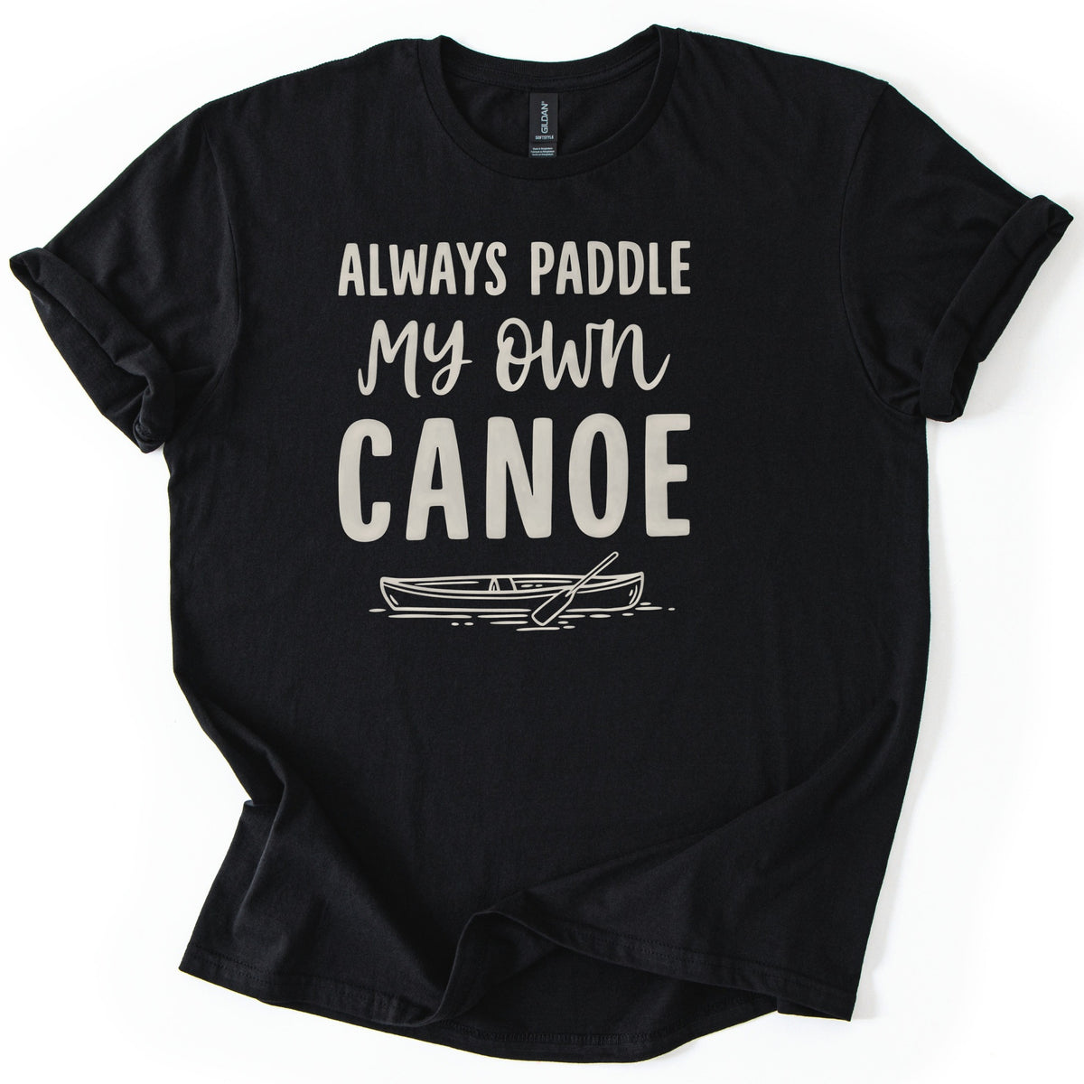 Paddle My Own Canoe Comfort Colors T-shirt in Black, laid flat. Cream text reads Always Paddle My Own Canoe above a line drawing of a canoe with a paddle on rippling water. The shirt has rolled sleeves and lies on a white surface.