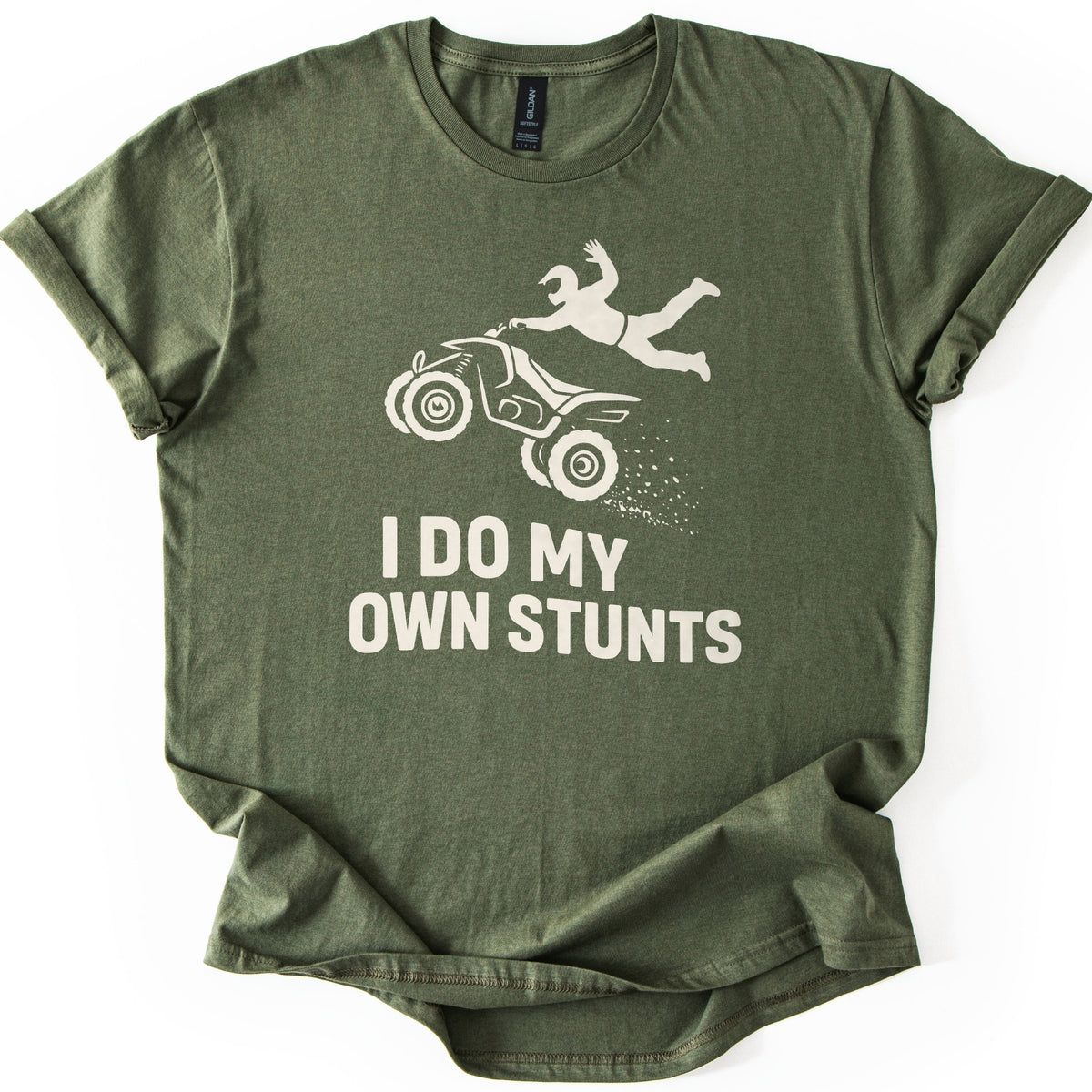ATV Stunts Comfort Colors T-shirt in Military Green, laid flat. A cream silhouette of a helmeted rider flying off the back of an ATV as it kicks up dirt, above the words I DO MY OWN STUNTS. The shirt lies on a white background with its sleeves cuffed.