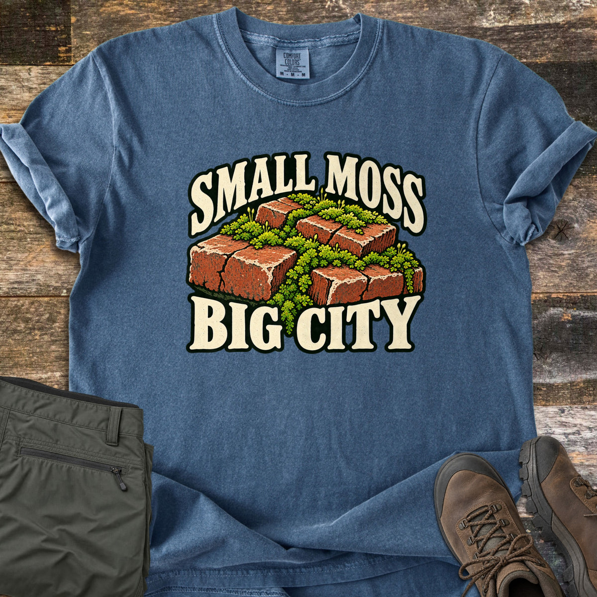Small Moss Big City Comfort Colors T-shirt in Denim, laid flat. Red bricks with bright green moss growing in the cracks, between the words Small Moss Big City. The shirt lies on weathered wood with olive green pants and brown hiking boots.