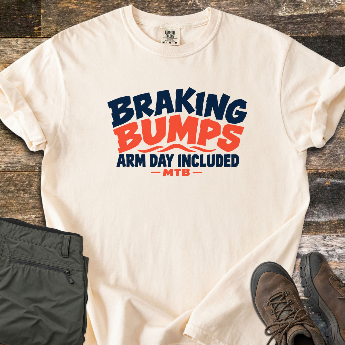 Braking Bumps Arm Day Included Comfort Colors T-shirt in Ivory, laid flat. Bold navy and orange text reads Braking Bumps, Arm Day Included, MTB, with wavy orange lines under Bumps. It rests on a wood floor beside olive cargo pants and brown hiking boots.