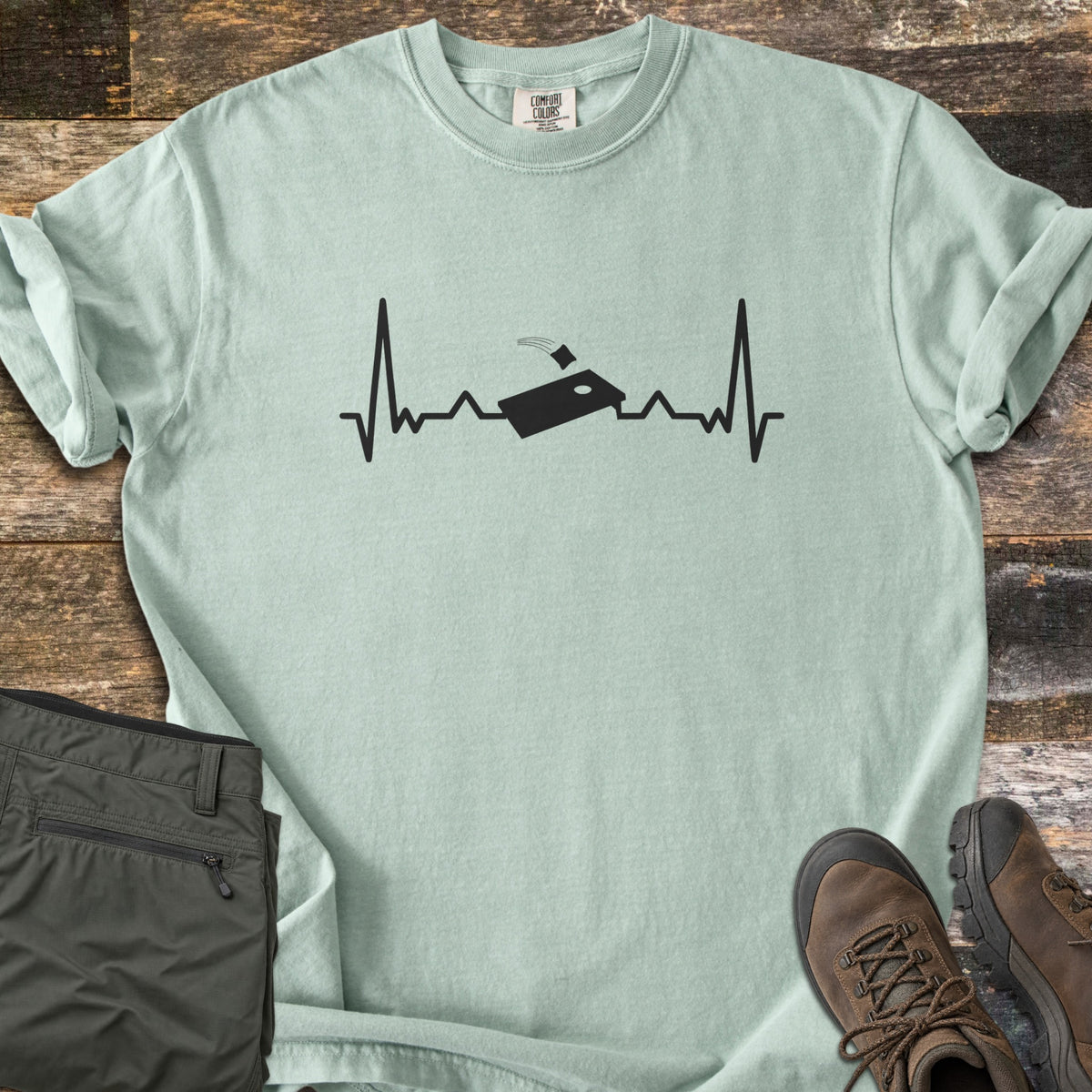 Corn Hole Comfort Colors T-shirt in Bay, laid flat. A black heartbeat line runs across the chest with a cornhole board at its center and a bean bag flying toward the hole. Olive pants and brown hiking boots sit beside it on weathered wood planks.