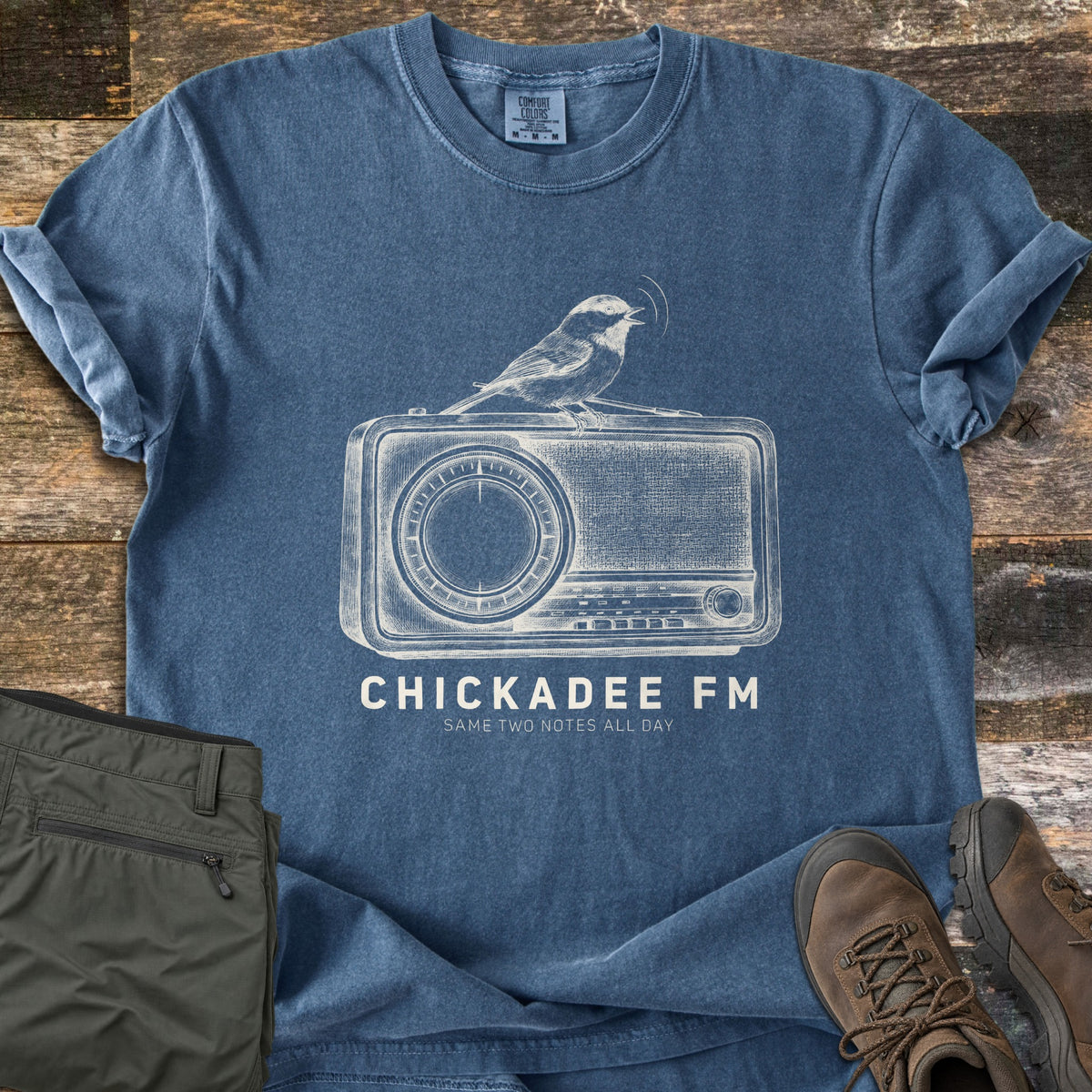 Chickadee FM Comfort Colors T-shirt in Denim, laid flat. A sketched chickadee sings atop a vintage radio above the words Chickadee FM and Same Two Notes All Day. Sleeves are rolled, on weathered wood beside folded olive hiking pants and brown hiking boots.