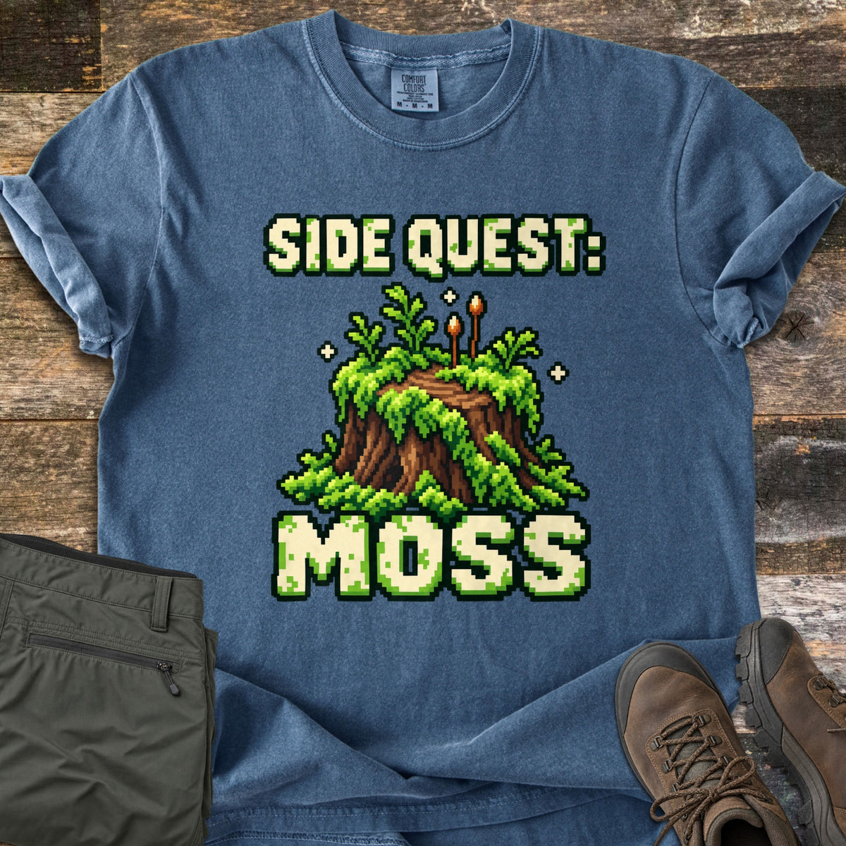 Side Quest Moss Comfort Colors T-shirt in Denim, laid flat. Pixel-art tree stump covered in green moss with ferns and two tiny mushrooms, between the words Side Quest: Moss. Olive cargo pants and brown hiking boots lie beside it on a weathered wood floor.