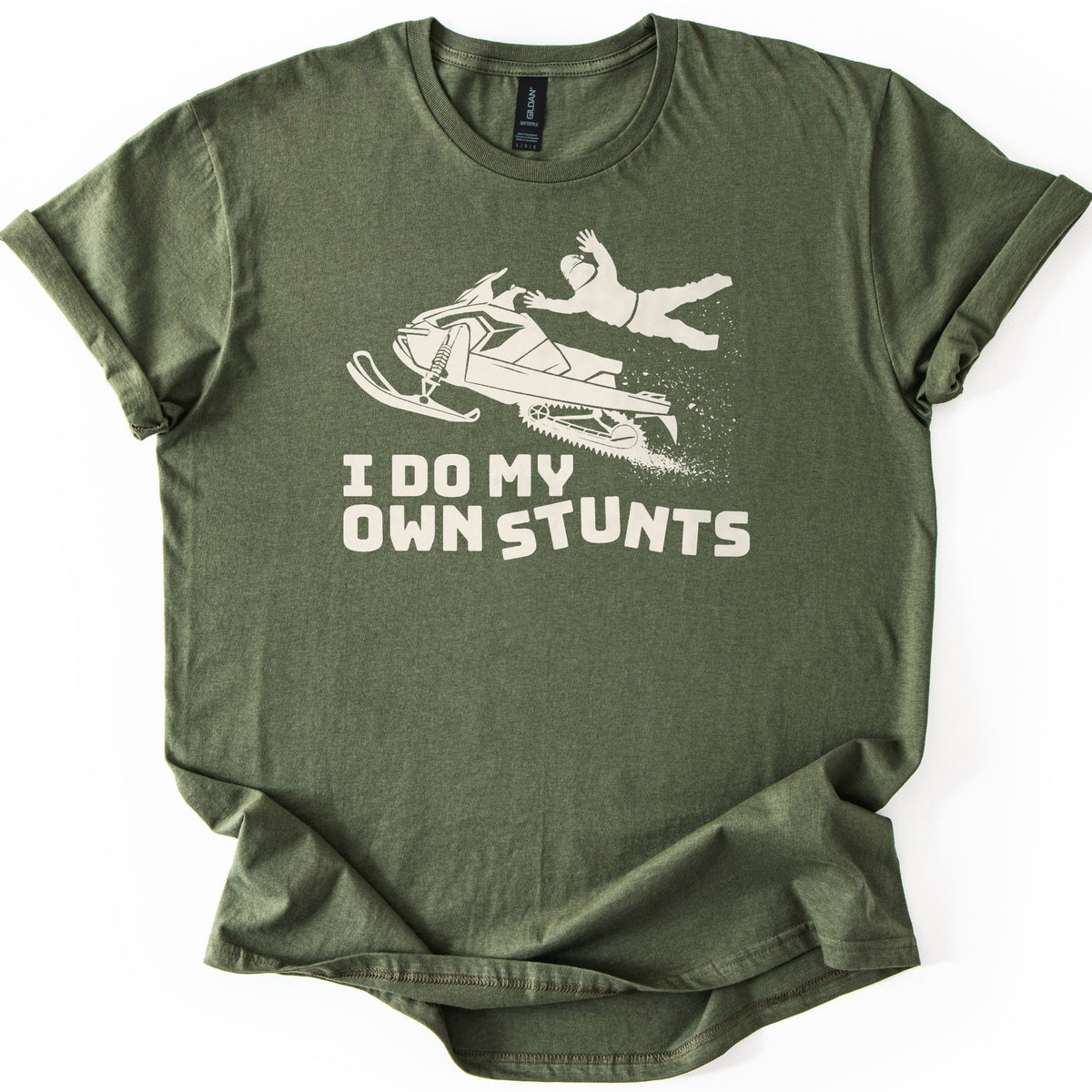 Snow Mobile Stunts Comfort Colors T-shirt in Military Green, laid flat. A cream silhouette of a snowmobile flying through the air, its rider thrown off behind it in a spray of snow, above the words I Do My Own Stunts. The shirt lies on a white background with rolled sleeves.