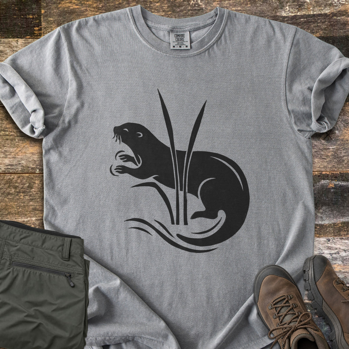 Otter In The Reeds Comfort Colors T-shirt in Grey, laid flat. A black silhouette of an otter with open mouth and raised paws curls behind two tall reeds above wavy water lines, no words. Folded olive hiking pants and brown hiking boots lie on weathered wood.