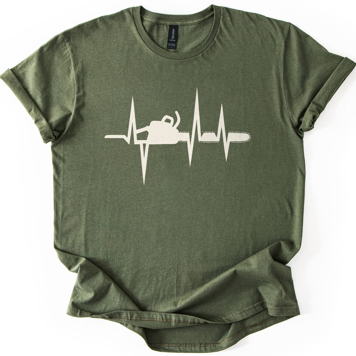 Chainsaw Heartbeat Comfort Colors T-shirt in Military Green, laid flat. A cream heartbeat line with sharp peaks runs across the chest, with a chainsaw silhouette set into the line, its bar and chain extending to the right. The sleeves are rolled on a white background.