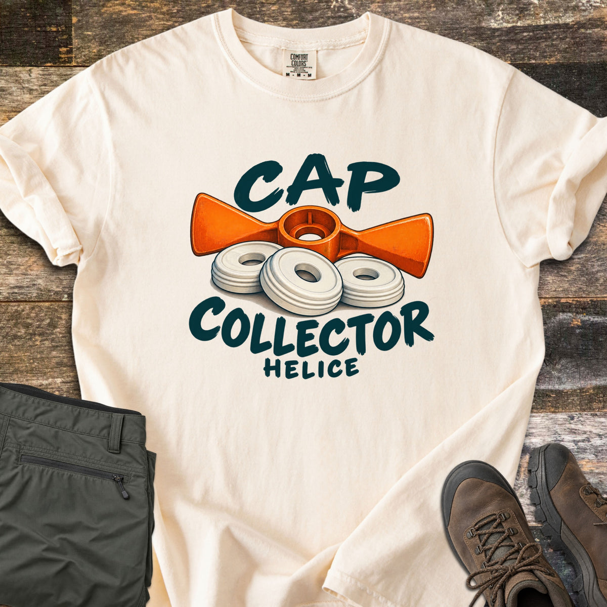 Cap Collector Comfort Colors T-shirt in Ivory, laid flat. An orange helice key and three white caps sit between the words Cap Collector, with Helice below. Sleeves are rolled; olive pants and brown hiking boots lie beside it on dark wood planks.