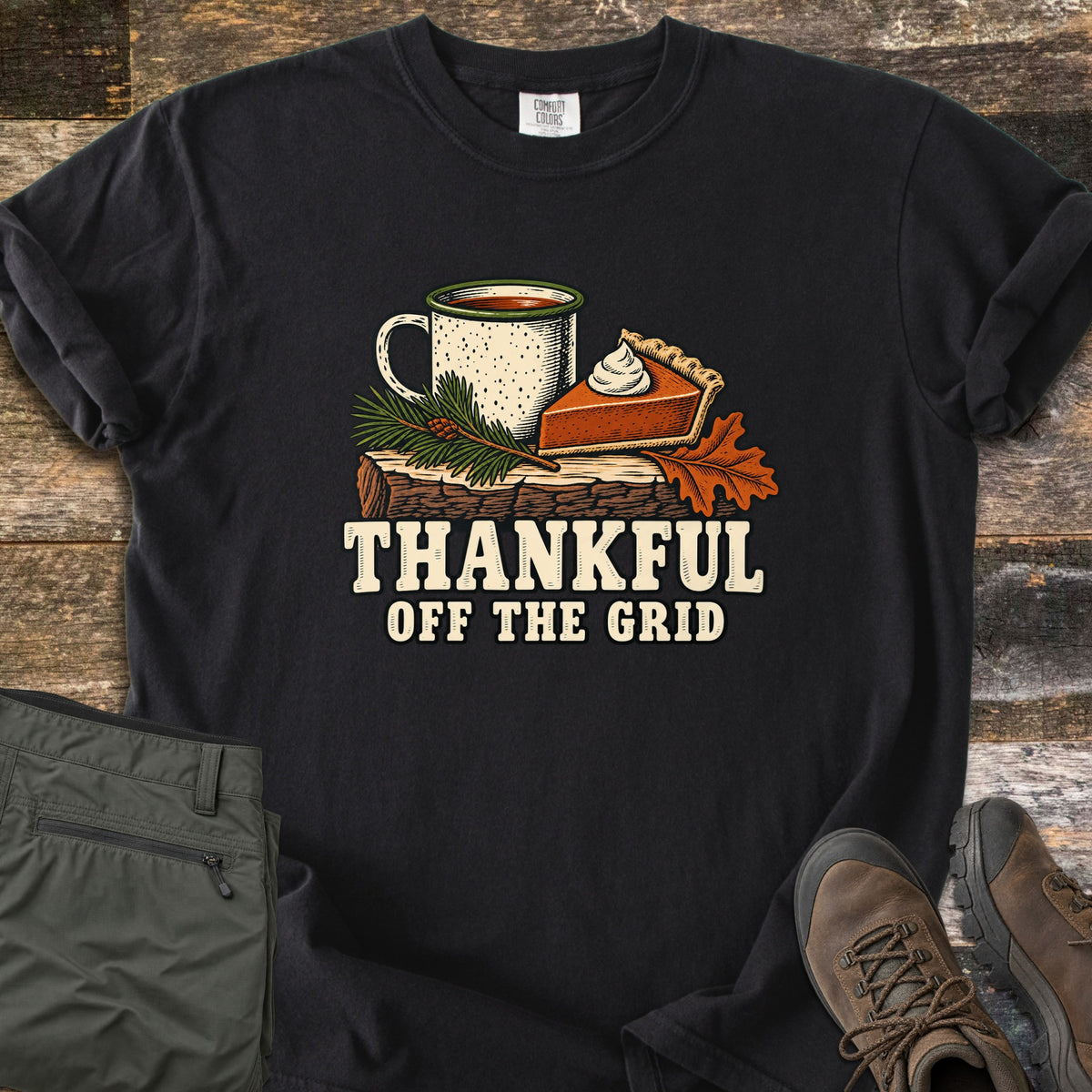 Thankful Off the Grid Comfort Colors T-shirt in Black, laid flat. A speckled enamel camp mug and a slice of pumpkin pie with whipped cream sit on a log with a pine sprig and an oak leaf above the words Thankful Off the Grid. Olive hiking pants and brown boots lie on weathered wood.