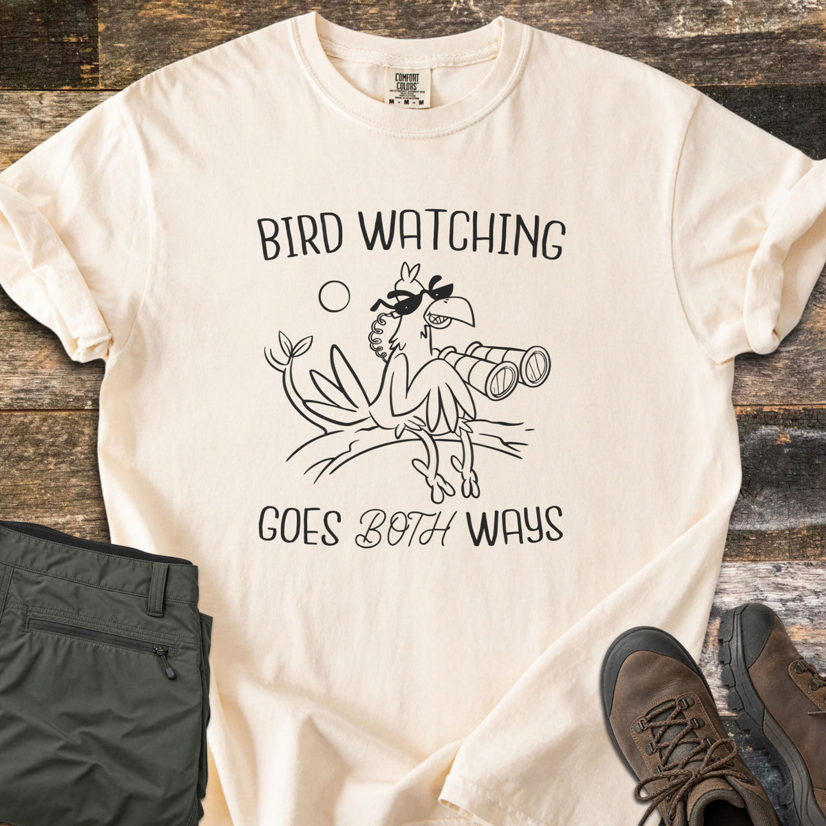 Birds Watch Back Birder Comfort Colors T-shirt in Ivory, laid flat. A cartoon bird in sunglasses perches on a branch beside binoculars between the words Bird Watching Goes Both Ways in black. Olive hiking pants and brown hiking boots sit beside it on weathered wood.