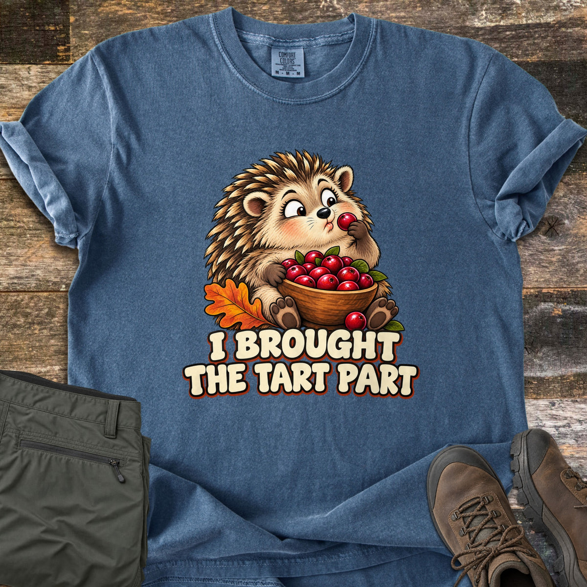 I Brought the Tart Part Comfort Colors T-shirt in Denim, laid flat. A cartoon hedgehog eats a cranberry while holding a bowl of cranberries beside an orange oak leaf, above the words I Brought the Tart Part. Shown on wood planks with olive cargo pants and brown hiking boots.