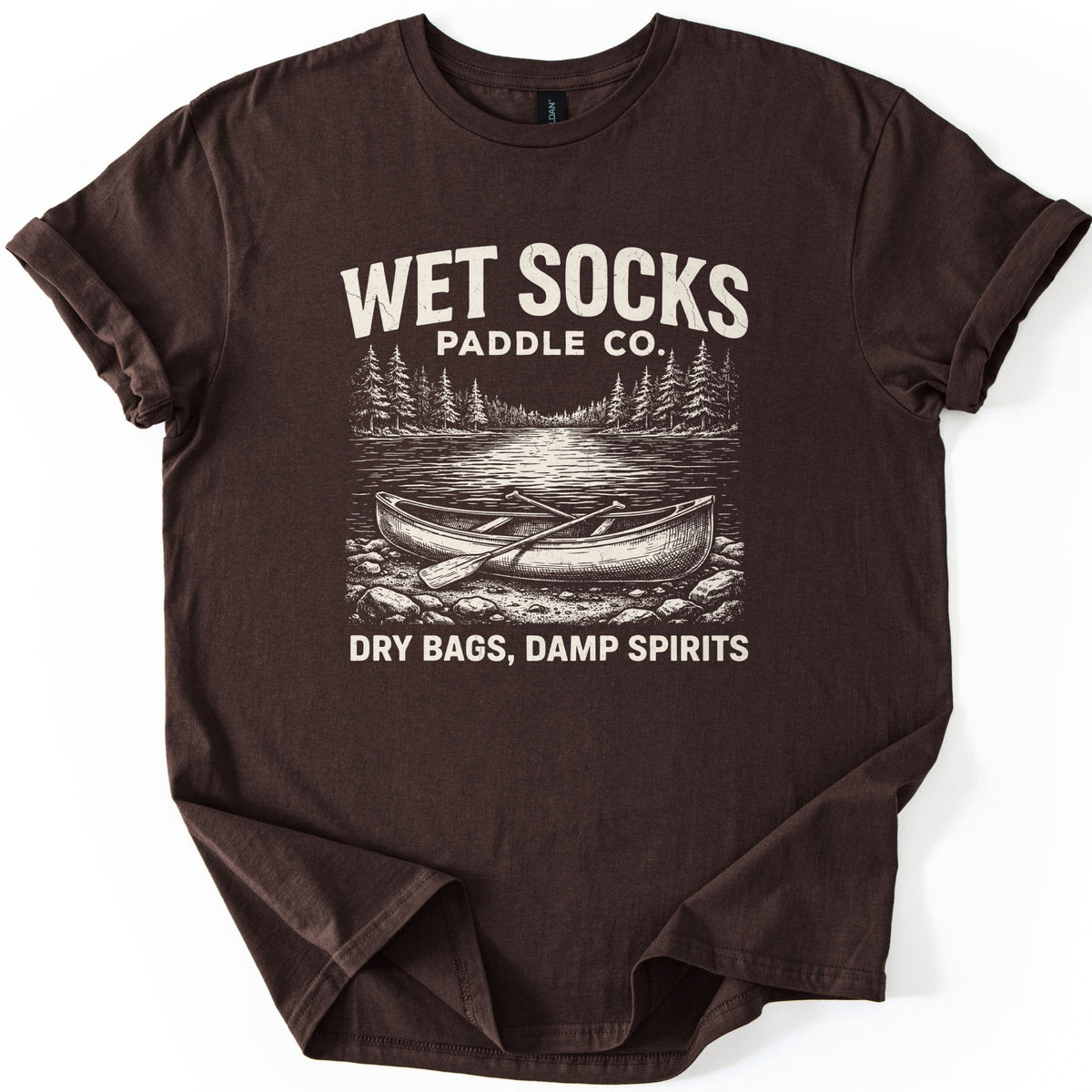Wet Socks Paddle Co Comfort Colors T-shirt in Dark Chocolate, laid flat. A cream etched drawing of a canoe with crossed paddles on a rocky shore, with a lake and pine trees behind, under the words Wet Socks Paddle Co. and above Dry Bags, Damp Spirits. White background.