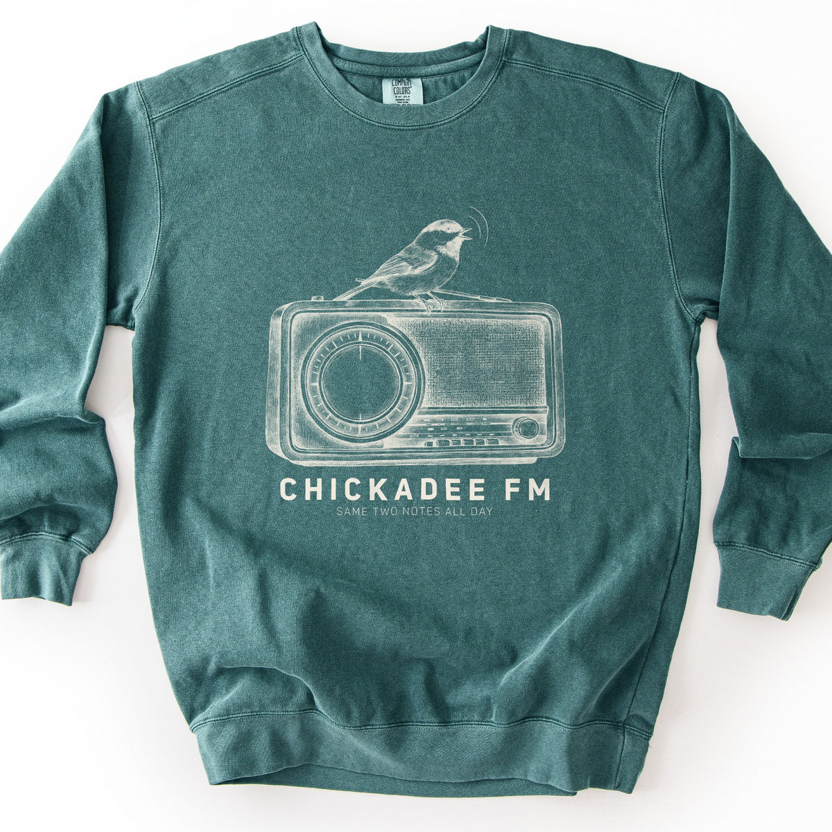 Chickadee FM Comfort Colors crewneck sweatshirt in Blue Spruce, laid flat. A vintage-style cream sketch of a chickadee singing atop an old radio, above the words CHICKADEE FM and SAME TWO NOTES ALL DAY. The sweatshirt lies on a plain white background.