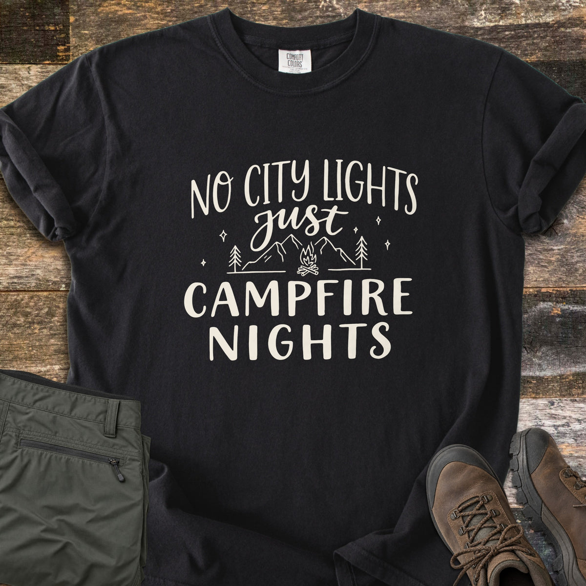Campfire Nights Comfort Colors T-shirt in Black, laid flat. Cream text reads No City Lights just Campfire Nights around line art of mountains, pine trees, stars and a campfire. Sleeves rolled, on weathered wood beside olive hiking pants and brown hiking boots.