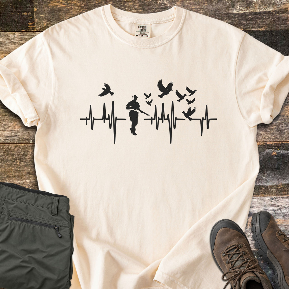 Bird Hunter Comfort Colors T-shirt in Ivory, laid flat. A black heartbeat line crosses the chest with a hunter silhouette in a cap holding a shotgun and birds flying up from the line. Folded olive pants and brown hiking boots lie beside it on weathered wood.