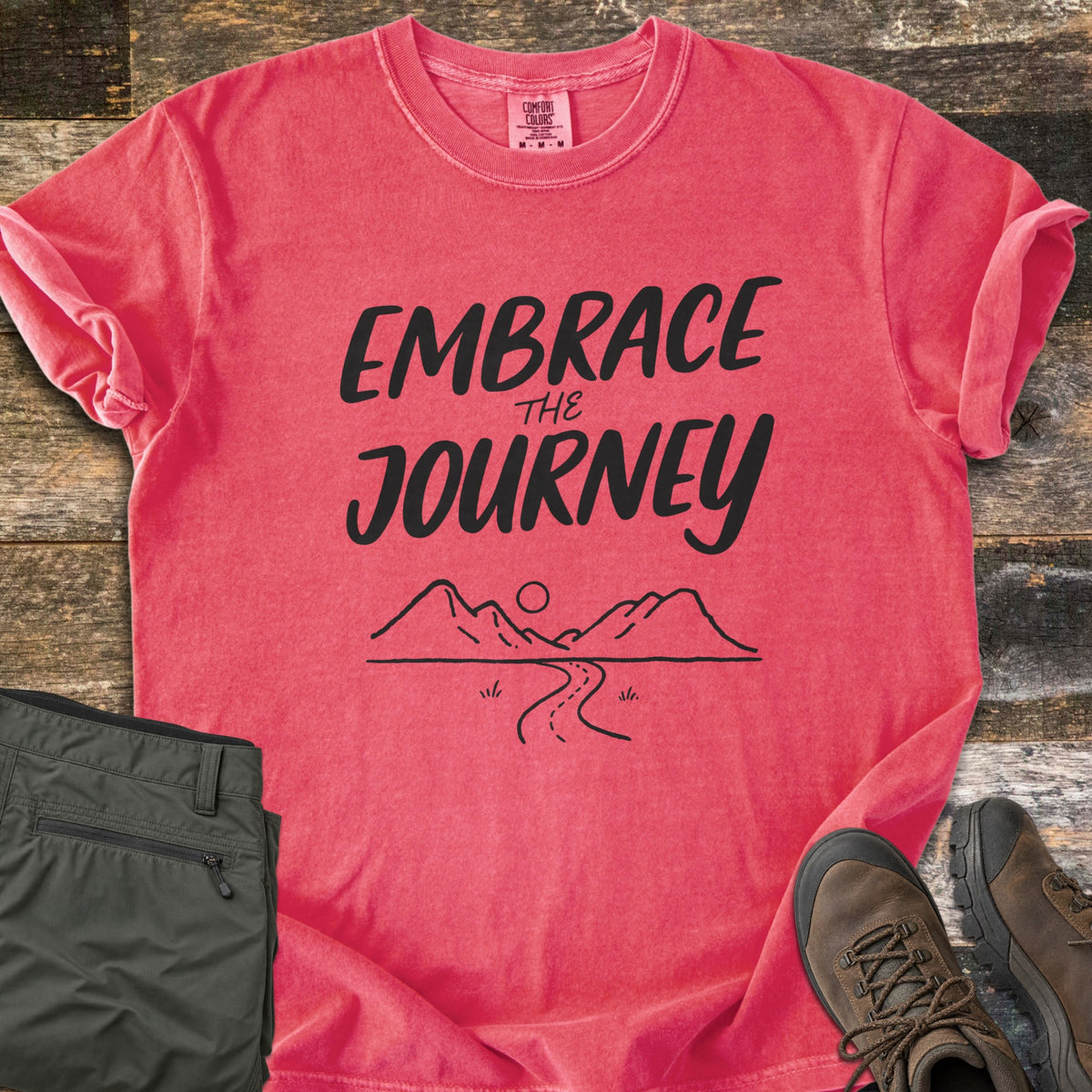 Embrace the Journey Comfort Colors T-shirt in Watermelon, laid flat. Black brush lettering reads Embrace the Journey above a line drawing of mountains, a sun, and a winding road. Sleeves cuffed on weathered wood beside folded green pants and brown hiking boots.