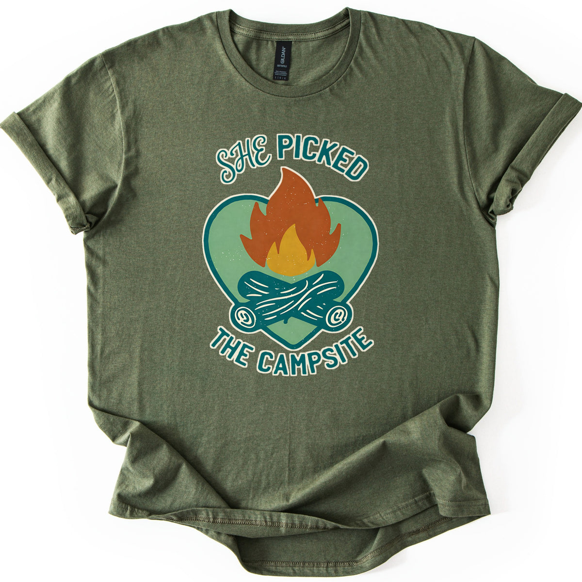 She Picked The Campsite Comfort Colors T-shirt in Military Green, laid flat. An orange and yellow campfire on crossed teal logs inside a sage green heart, with the words SHE PICKED above and THE CAMPSITE below. The shirt lies on a white surface with its sleeves cuffed.