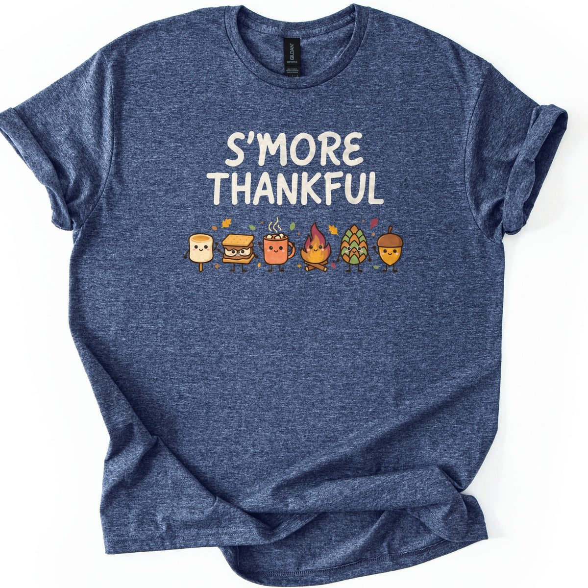 Smore Thankful Comfort Colors T-shirt in Heather Navy, laid flat. Cream letters read S'MORE THANKFUL above a row of smiling cartoon characters: a marshmallow, a s'more, a cocoa mug, a campfire, a pinecone and an acorn among autumn leaves. Sleeves rolled, on white.