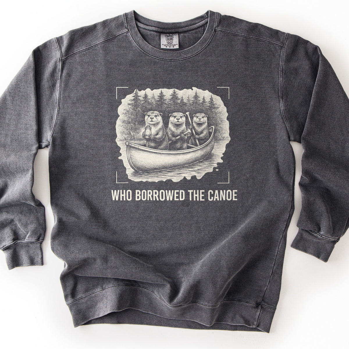 Who Borrowed The Canoe Comfort Colors crewneck sweatshirt in Pepper, laid flat. A cream sketch of three otters in a canoe on a lake, one holding a lantern and two holding paddles, pine trees behind, framed by corner brackets. Below it WHO BORROWED THE CANOE in cream. Laid on white.