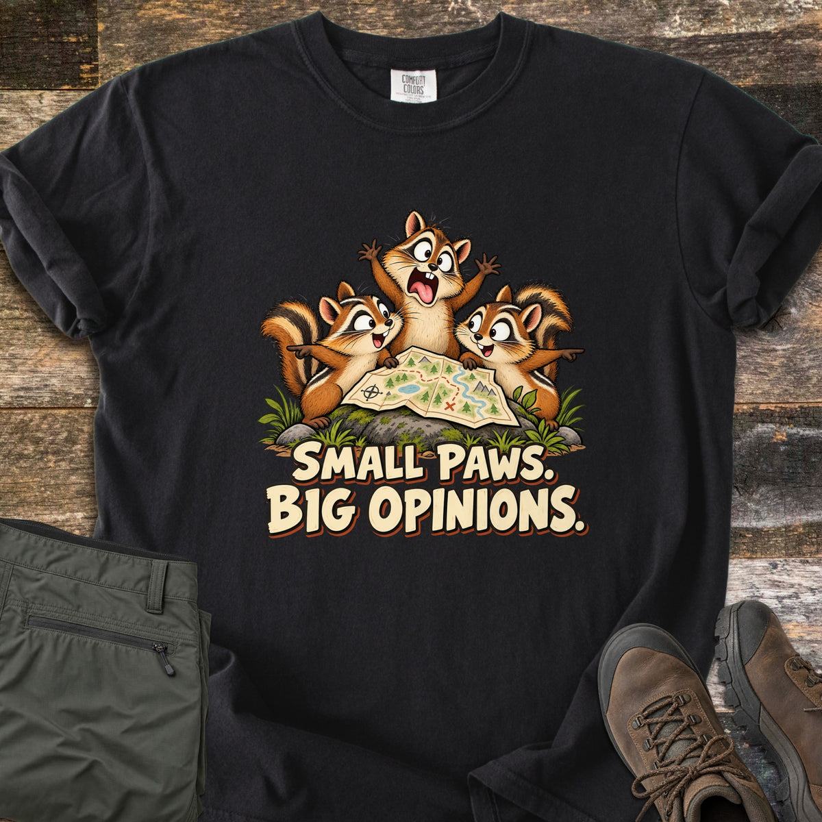 Small Paws. Big Opinions. Comfort Colors T-shirt in Black, laid flat. Three cartoon chipmunks argue over a trail map on a mossy rock, the middle one yelling with arms raised, above the words SMALL PAWS. BIG OPINIONS. Olive hiking pants and brown boots sit beside it on weathered wood.