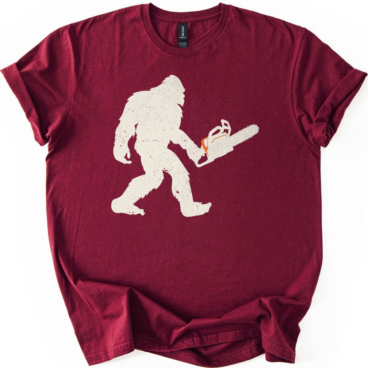Lumber Squatch Comfort Colors T-shirt in Maroon, laid flat. A distressed cream silhouette of Bigfoot walking mid-stride while holding a chainsaw with an orange accent, with no words printed. The shirt has cuffed sleeves and lies on a white background.