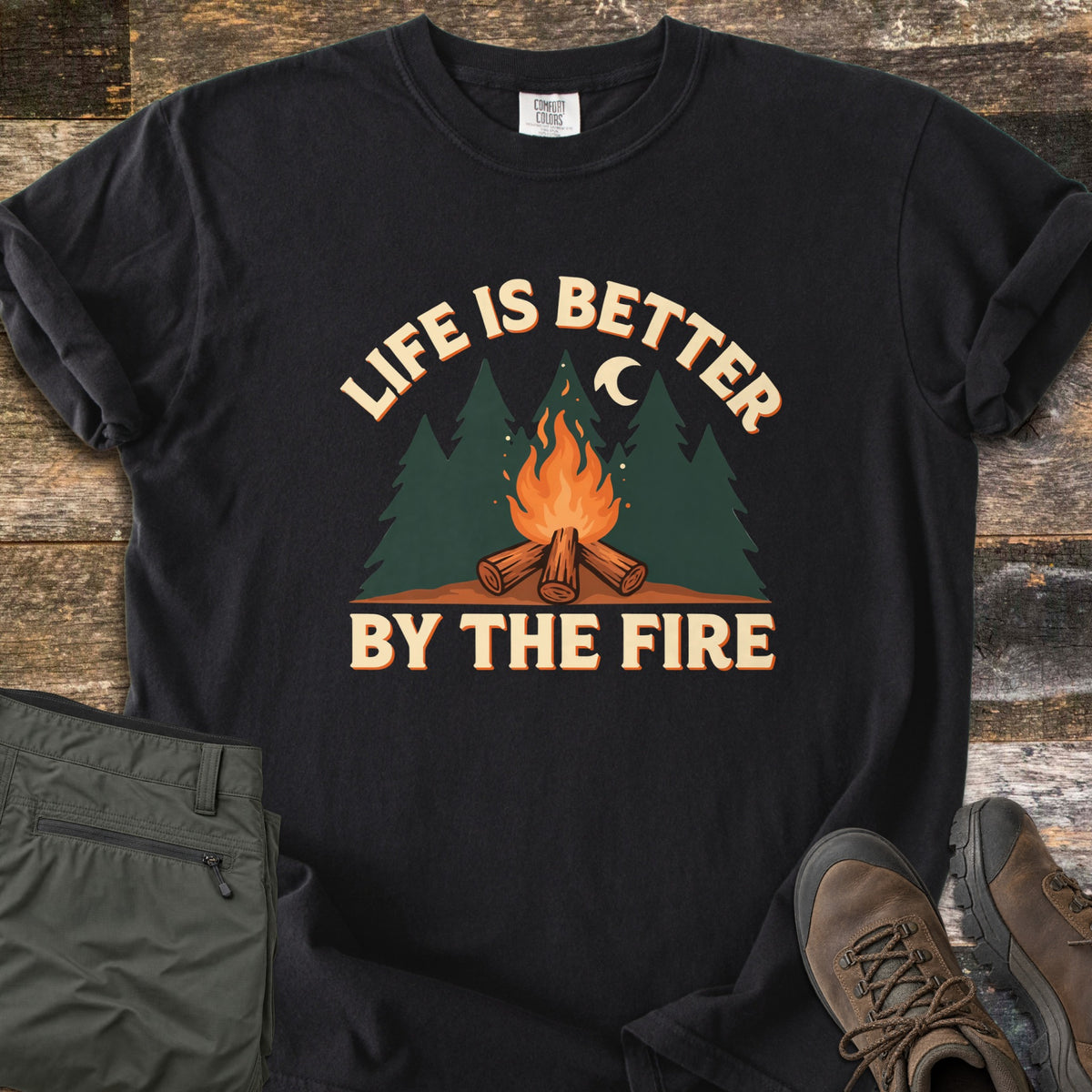 Life Is Better By The Fire Comfort Colors T-shirt in Black, laid flat. Arched cream text reads Life Is Better over dark green pine trees, a crescent moon and a campfire on crossed logs, with By The Fire below. Folded olive pants and brown hiking boots sit on weathered wood.