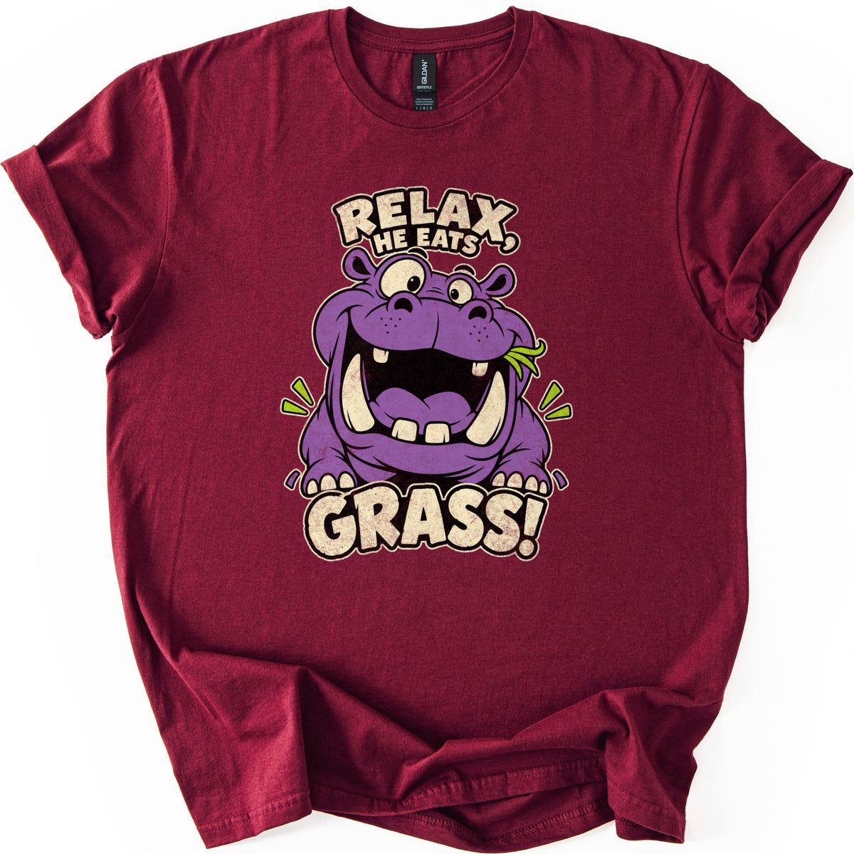 Relax He Eats Grass Comfort Colors T-shirt in Maroon, laid flat. A grinning purple cartoon hippo with big tusks and a blade of grass in its mouth sits between the words RELAX, HE EATS and GRASS! in cream letters. The shirt lies on white with rolled sleeves.