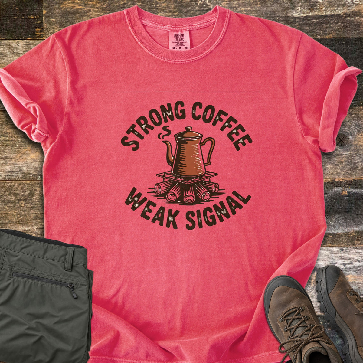 Strong Coffee Weak Signal Comfort Colors T-shirt in Watermelon, laid flat. A steaming copper coffee pot sits on a grate over campfire logs, arched by the words Strong Coffee above and Weak Signal below. Folded olive hiking pants and brown hiking boots lie on weathered wood planks.