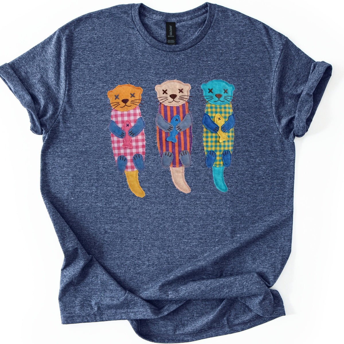 Patchwork Otter Comfort Colors T-shirt in Heather Navy, laid flat. Three appliqué otters with X-stitched eyes float side by side holding fish: orange in pink gingham, beige in purple and orange stripes, turquoise in yellow gingham. No words. Rolled sleeves on a white background.
