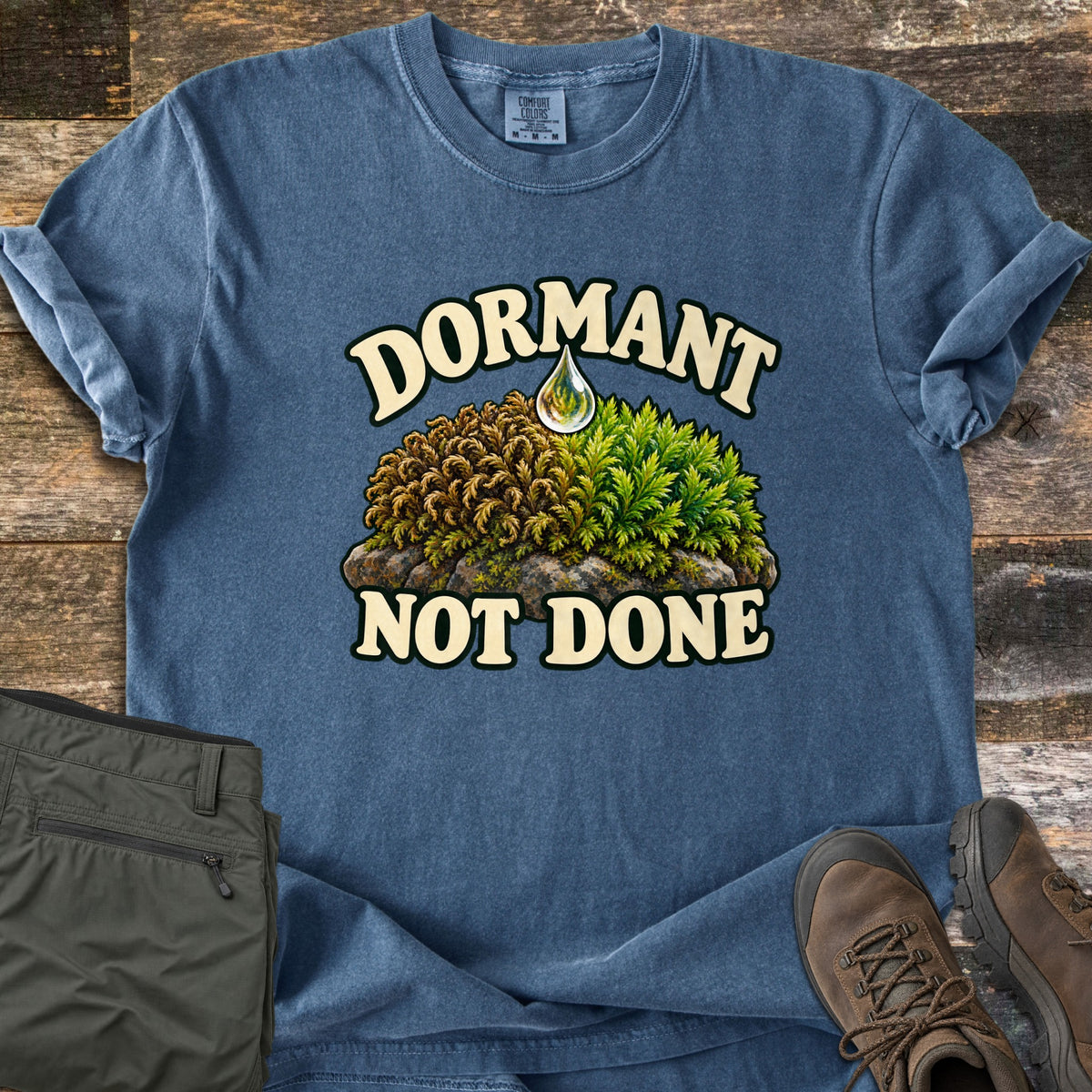 Dormant Not Done Comfort Colors T-shirt in Denim, laid flat. A mound of moss on a rock, brown and dry on the left and green on the right, sits under a water droplet between the words Dormant Not Done. Olive hiking pants and brown hiking boots lie beside it on weathered wood.
