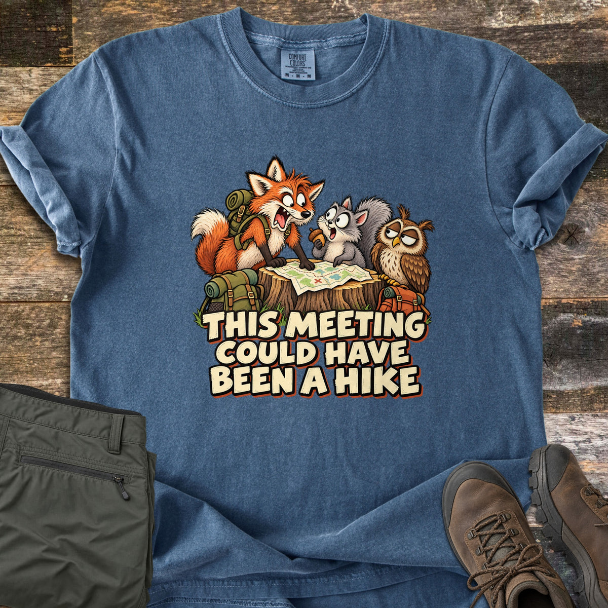 This Meeting Could Have Been a Hike Comfort Colors T-shirt in Denim, laid flat. A cartoon fox with a backpack, a gray squirrel and an owl study a map on a tree stump above the words This Meeting Could Have Been a Hike. It lies on wood planks beside olive hiking pants and brown boots.