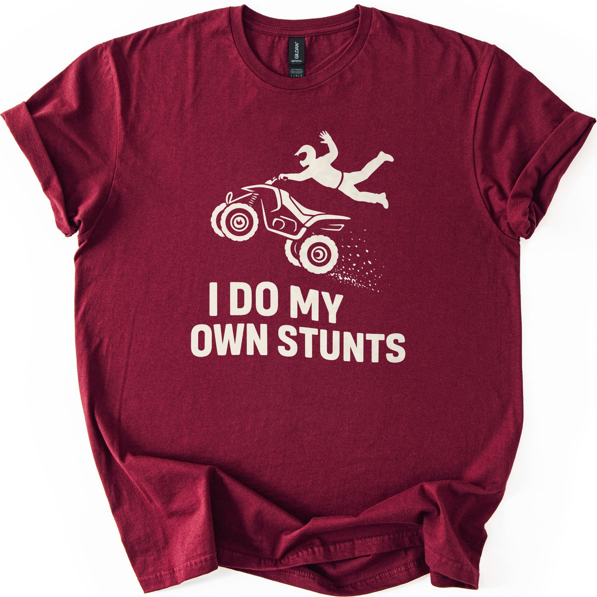 ATV Stunts Comfort Colors T-shirt in Maroon, laid flat. A cream silhouette of a helmeted rider flying off the back of an ATV as it kicks up dirt, above the words I DO MY OWN STUNTS. The shirt lies on a white background with its sleeves cuffed.