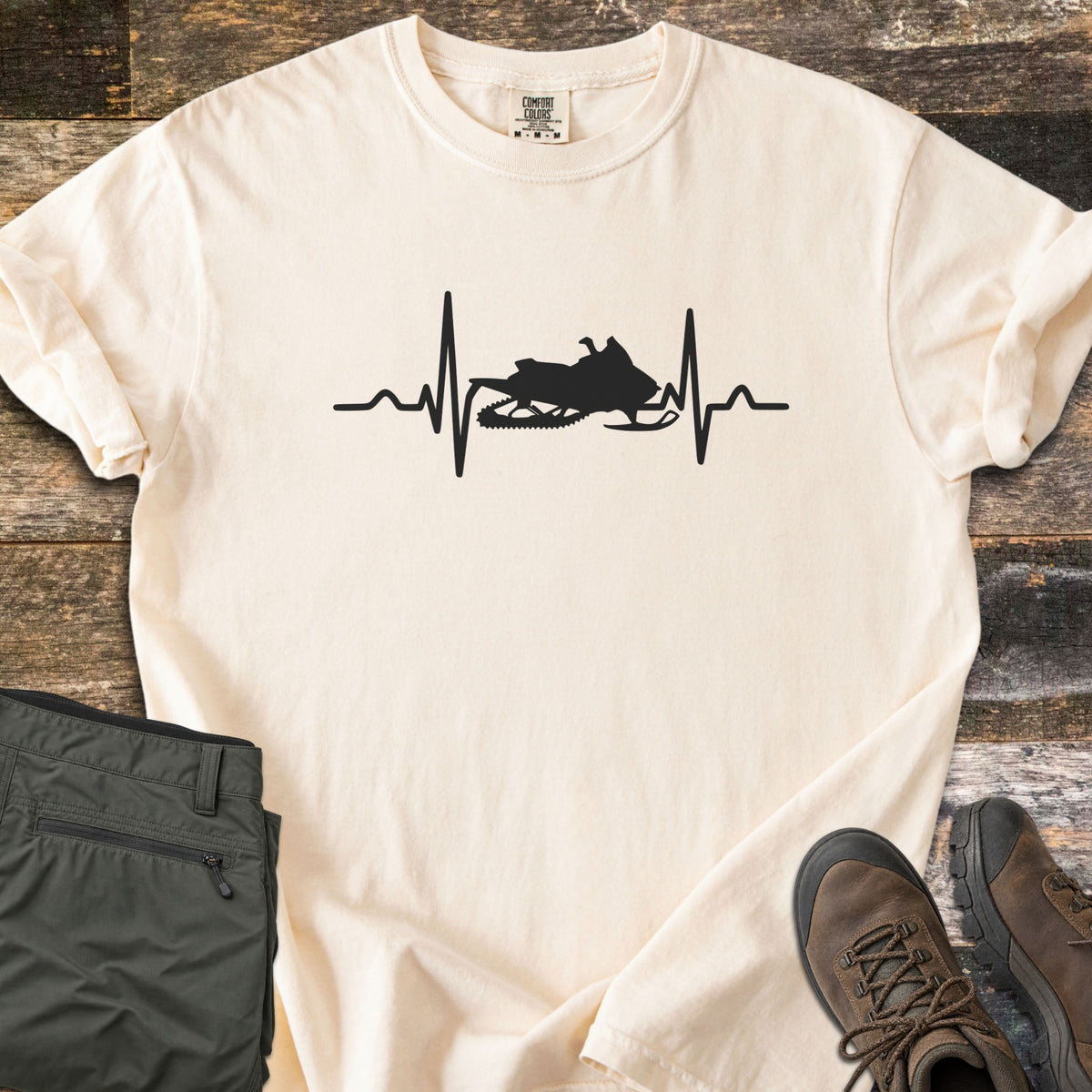 Snowmobile Heartbeat Comfort Colors T-shirt in Ivory, laid flat. A black heartbeat line runs across the chest with a snowmobile silhouette in its center, with no words. Sleeves are rolled, on weathered wood planks beside folded olive pants and brown hiking boots.