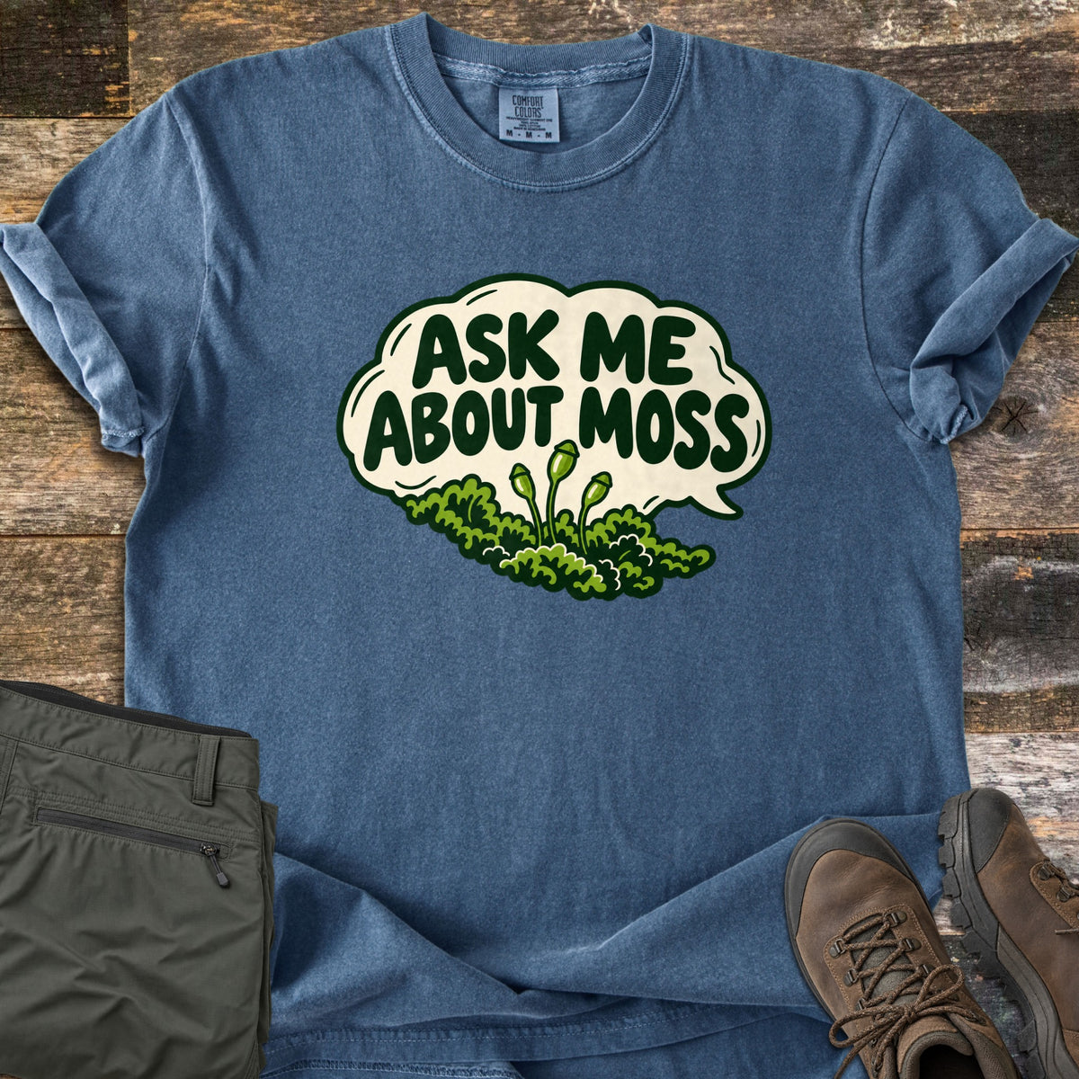 Ask Me About Moss Comfort Colors T-shirt in Denim, laid flat. A cream speech bubble reads Ask Me About Moss in dark green letters above a clump of green moss with sprouting capsules. Sleeves are cuffed on weathered wood beside olive hiking pants and brown hiking boots.