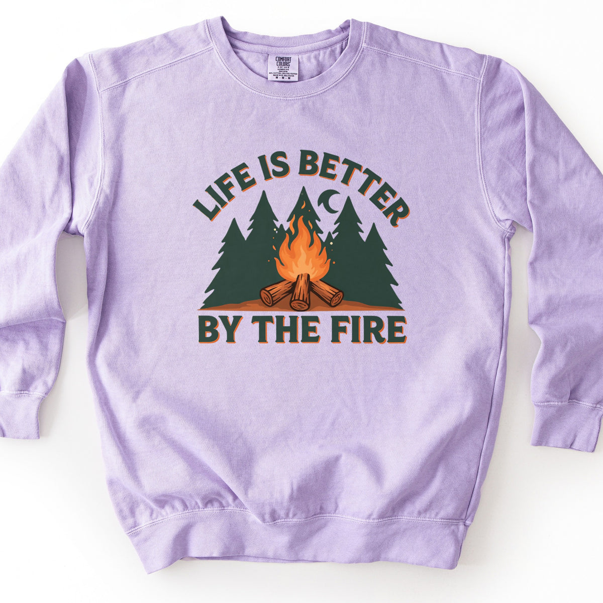 Life Is Better By The Fire Comfort Colors crewneck sweatshirt in Orchid, laid flat. Dark green letters with an orange shadow arch Life Is Better over dark green pine trees, a crescent moon and a campfire of crossed logs, with By The Fire below. It lies on a white surface.