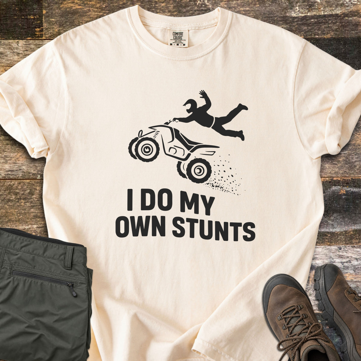 ATV Stunts Comfort Colors T-shirt in Ivory, laid flat. A black silhouette of a helmeted rider flying off a wheelie-popping ATV kicking up dirt, above the words I Do My Own Stunts. Olive cargo pants and brown hiking boots sit beside it on weathered wood.