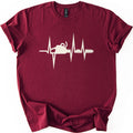 Chainsaw Heartbeat Comfort Colors T-shirt in Maroon, laid flat. A cream heartbeat line with sharp peaks runs across the chest, with a chainsaw silhouette set into the line, its bar and chain extending to the right. The sleeves are rolled on a white background.