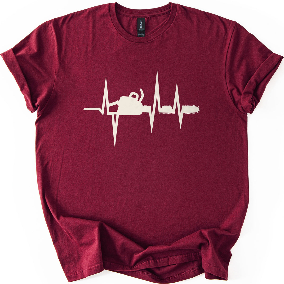 Chainsaw Heartbeat Comfort Colors T-shirt in Maroon, laid flat. A cream heartbeat line with sharp peaks runs across the chest, with a chainsaw silhouette set into the line, its bar and chain extending to the right. The sleeves are rolled on a white background.