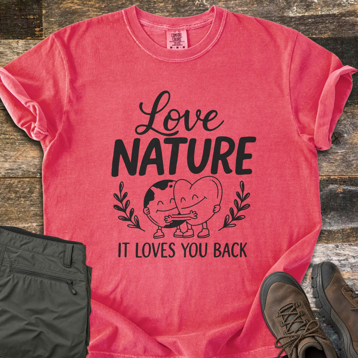 Nature Loves You Back Comfort Colors T-shirt in Watermelon, laid flat. Black print reads Love Nature above a smiling cartoon Earth and heart hugging between leafy sprigs, with It Loves You Back below. It lies on weathered wood beside olive hiking pants and brown boots.