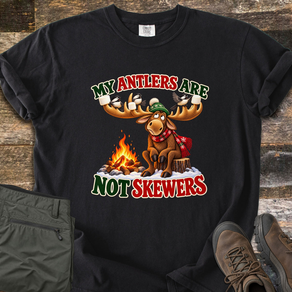 My Antlers Are Not Skewers Comfort Colors T-shirt in Black, laid flat. A cartoon moose in a green knit hat and red scarf sits on a stump by a campfire as chickadees on its antlers hold marshmallows, with the words My Antlers Are Not Skewers. Olive pants and brown boots lie on weathered wood.