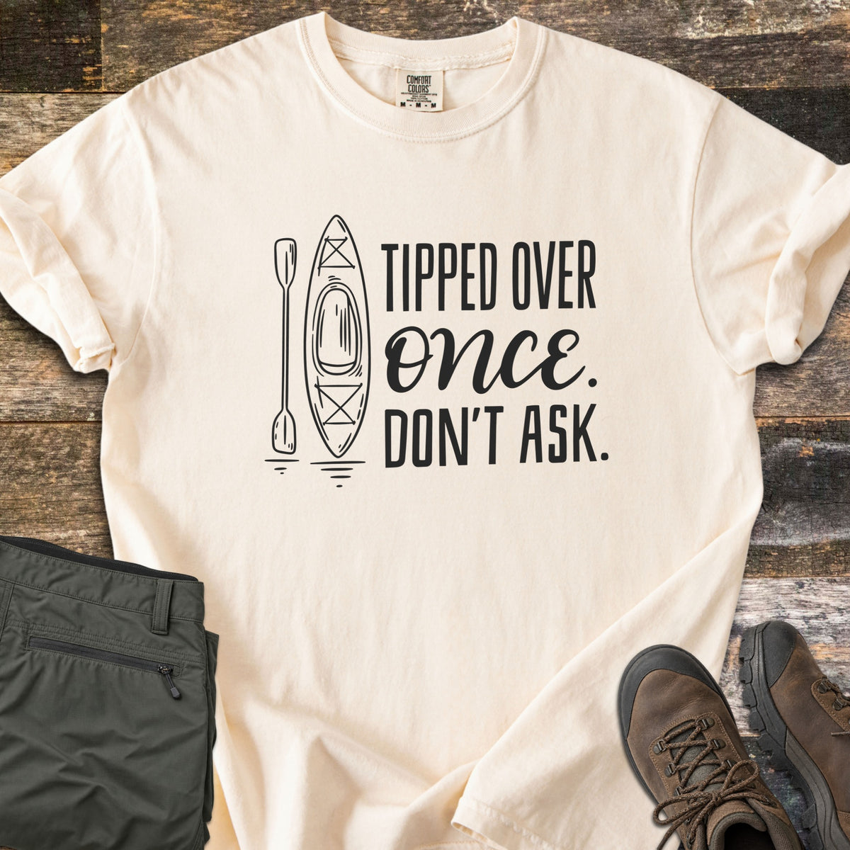 Tipped Over Once. Don't Ask. Comfort Colors T-shirt in Ivory, laid flat. A black line drawing of a kayak and paddle sits beside the words Tipped Over Once. Don't Ask., with Once in script. Sleeves rolled, on weathered wood with folded olive hiking pants and brown hiking boots.
