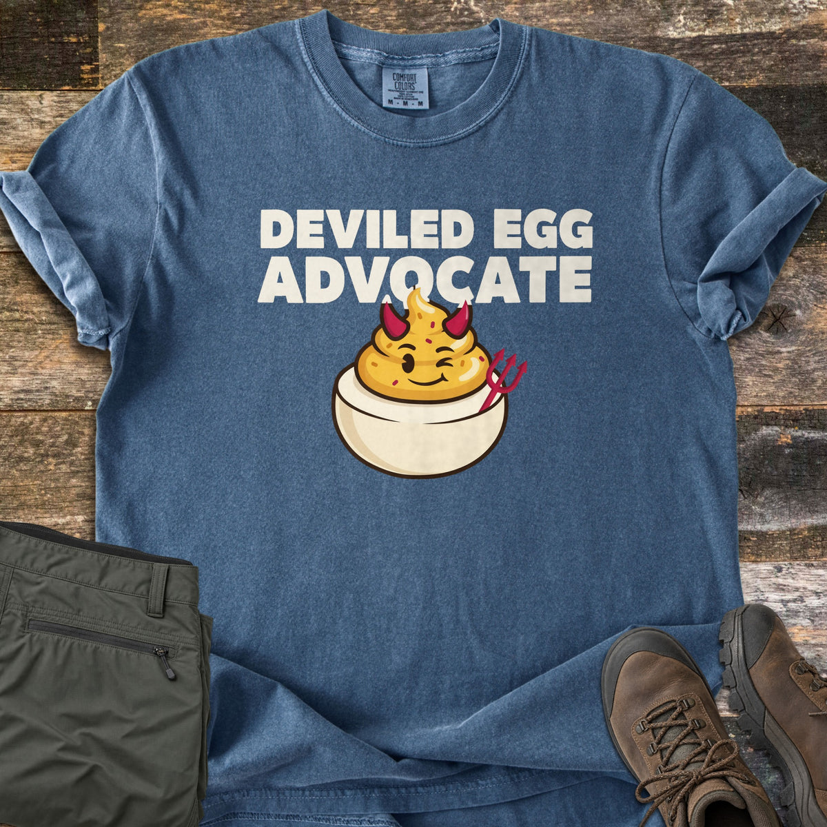 Deviled Egg Advocate Comfort Colors T-shirt in Denim, laid flat. White text reads DEVILED EGG ADVOCATE above a winking cartoon deviled egg with red devil horns and a pitchfork. It lies on weathered wood beside folded olive green pants and brown hiking boots.