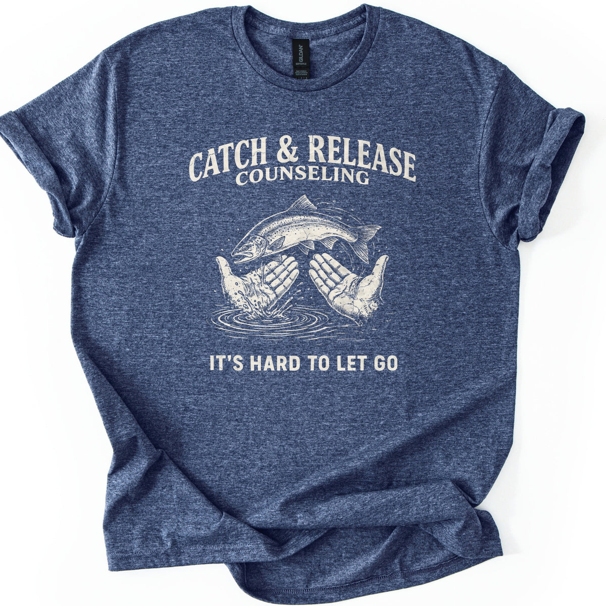 Catch & Release Counseling Comfort Colors T-shirt in Heather Navy, laid flat. Cream print reads CATCH & RELEASE COUNSELING above an etched trout leaping from two open hands over rippling water, with IT'S HARD TO LET GO below. Sleeves are cuffed on a white background.
