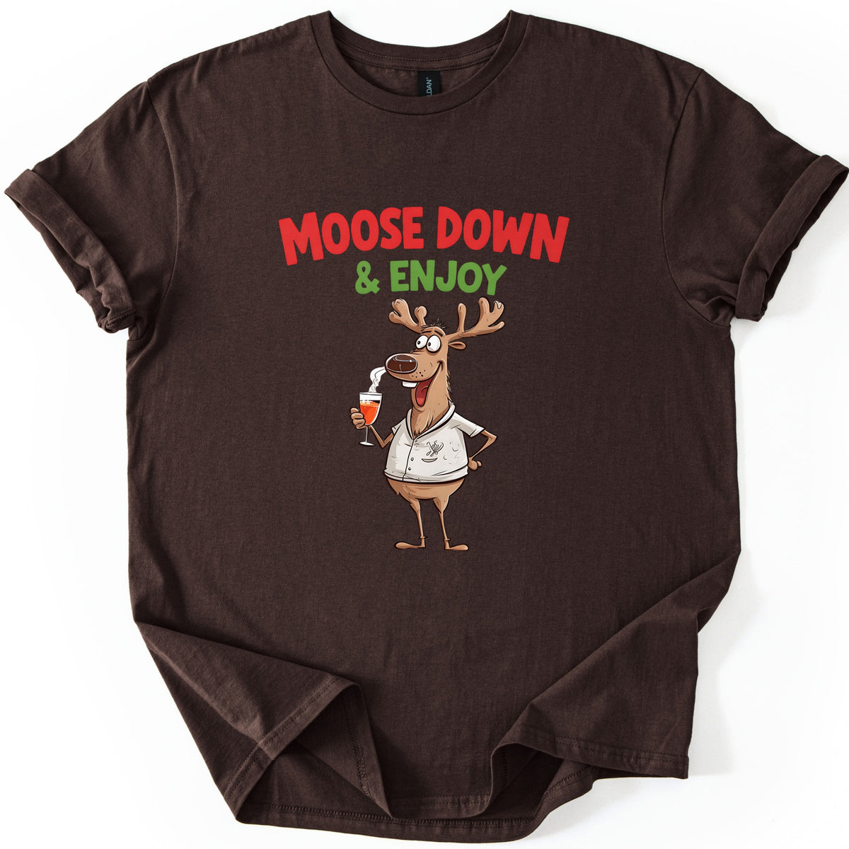 Moose Down And Enjoy Comfort Colors T-shirt in Dark Chocolate, laid flat. Red words Moose Down above green & Enjoy, over a grinning cartoon moose in a white short-sleeve shirt holding a glass of orange drink, hand on hip. Sleeves are cuffed on a white background.