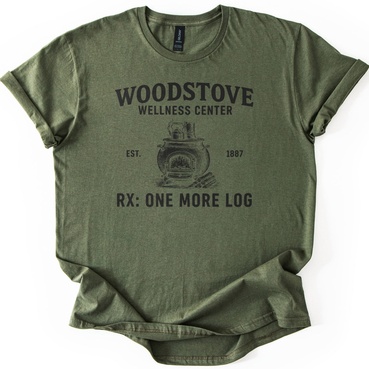 Woodstove Wellness Center Comfort Colors T-shirt in Military Green, laid flat. A black sketch of a round woodstove with a kettle on top, flames in its open door and stacked logs beside it, under Woodstove Wellness Center, Est. 1887, and Rx: One More Log. Sleeves rolled, on white.