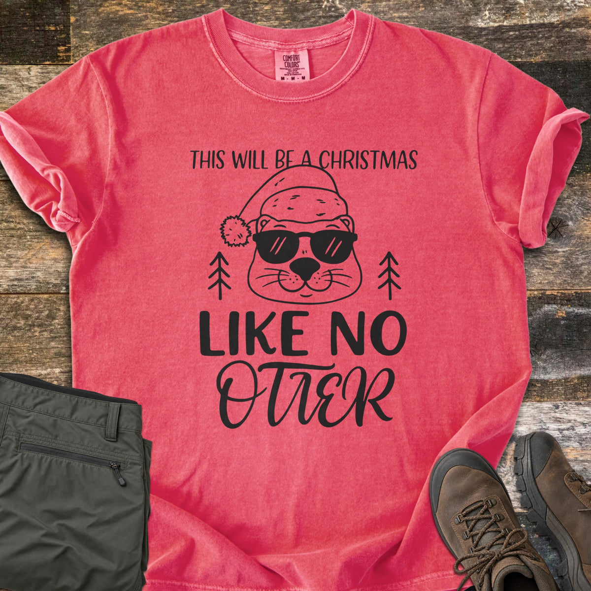 Christmas Like No Otter Comfort Colors T-shirt in Watermelon, laid flat. A cartoon otter in a Santa hat and sunglasses sits between two pine trees, with This Will Be a Christmas above and Like No Otter below, in black. Olive cargo pants and brown hiking boots lie on weathered wood.