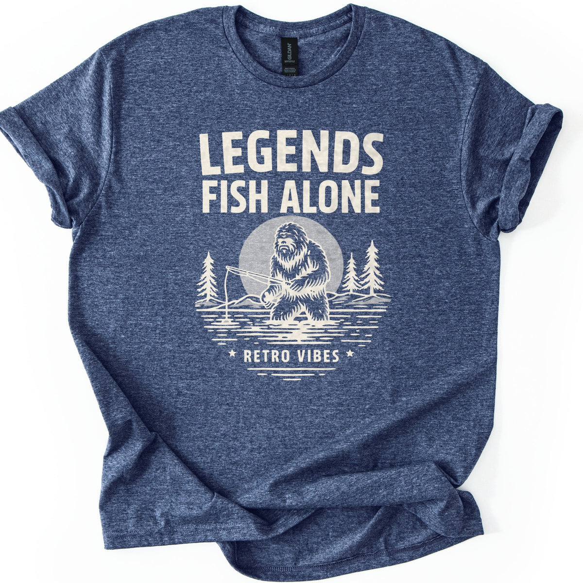 Legends Fish Alone Bigfoot Comfort Colors T-shirt in Heather Navy, laid flat. Cream print of Bigfoot fishing knee-deep in a lake before a light gray moon and pine trees, under the words Legends Fish Alone and above Retro Vibes between two stars. Sleeves cuffed, on a white background.